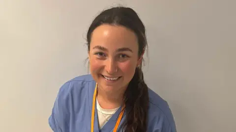 A headshot of a woman who is wearing pale blue scrubs. She has dark brown hair which is tied up in a ponytail and is smiling into the camera. She has an orange lanyard around her neck and a nose ring. 