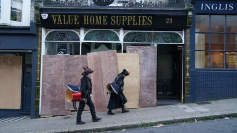 PA Medium Two people in black clothes walking past a board shop. Above the store is a black sign that says VALUE HOME SUPPLYS in gold letters