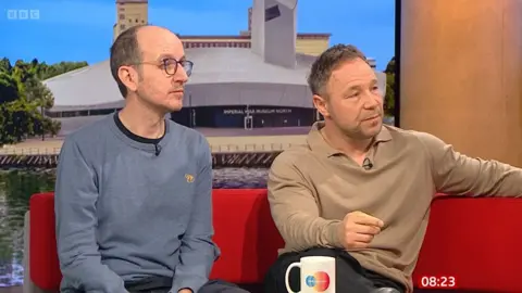 Jack Thorne (left) and Stephen Graham sit on a red sofa in the BBC. Jack wears a blue sweater, he has short hair and wears black glasses. Stephen has a light brown collar sweater.