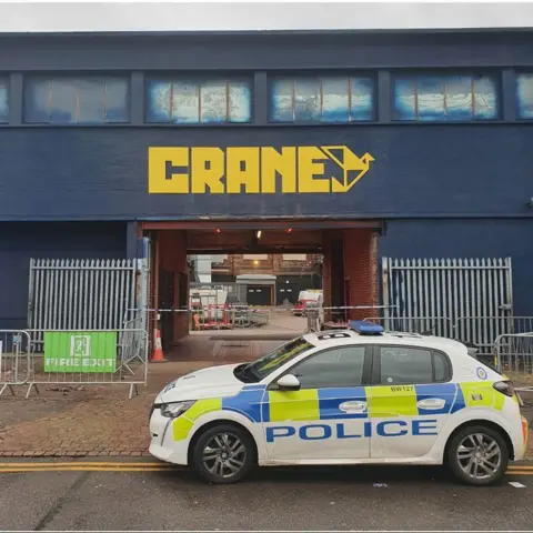 West Midlands Police A police car parked outside a dark blue building with the word "Crane" in yellow lettering above an entrance.