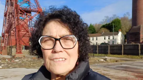 BBC News Sian woman looks towards the camera with black hair and black framed glasses. She wears a black coat and is on former mining land. 