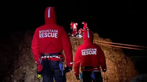 Two members of a mountain rescue team look on at their colleagues on a rocky hill carrying out a rescue operation.