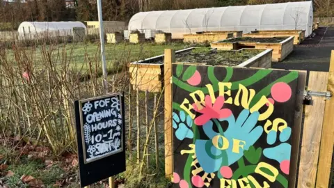 A large community garden. It has a multicoloured hand painted sign that says 'friends of the field' on the front gate. Another black and white sign says 'FOF opening hours 10-1pm Sat'. There are two large white polytunnels and wooden raised beds in the garden. 