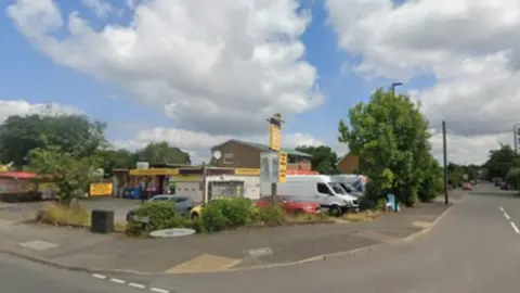 Two roads join with what appears to a car wash on the corner of the junction. Some vans and cars are parked on the forecourt with trees lining the road.