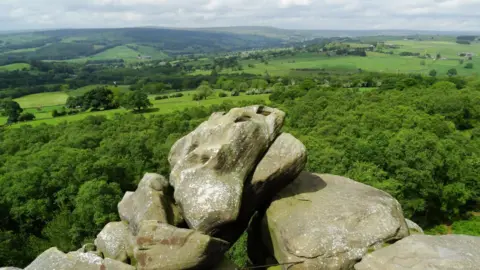 In the foreground is a carefully balance rock formation. Behind is a view of rolling, green fields, trees and moors.