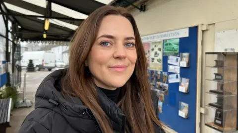 A woman with long dark hair wearing a black coat, standing at the entrance to Hythe Pier with the ferry information board in the background