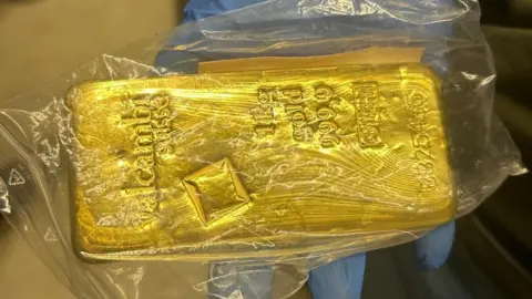 A person wearing blue gloves holding a bar of gold in a plastic bag. The bar has numbers, writing and shapes on it. 