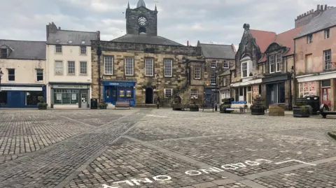 The cobbles of Alnwick's market place stetch out towards some historic looking buildings which fringe two corners of the photograph.