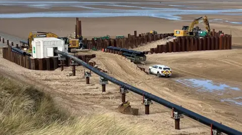 This image shows Talacre Beach, There's construction works happening in the background, The long line of structures now have a black pipe resting on top of them. There's long grass and sand around the construction site.