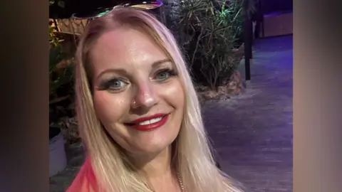 Head-and-shoulders of Michaela Wilson who has long, straight blonde hair, blue eyes, red lipstick and has a pair of sunglasses on top of her head. 