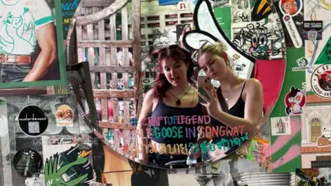 Gwen Shepherd Mia and Gwen taking a selfie of themselves in a mirror. The mirror is on a wall which is covered with various pictures and slogans.