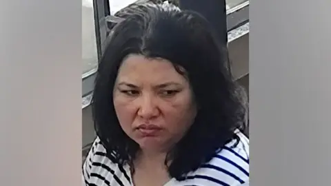 A woman with dark hair and a serious expression looks down. She wears a striped black and white shirt.