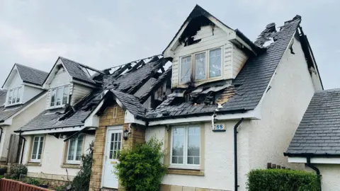 A white detached house with a burnt in roof. The windows are smashed and there are holes in the roof.