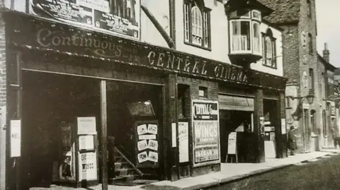 Museum of Cambridge Black and white photo of two storeys of an art-deco cinema in a building that is clearly older than the cinema which opened in 1930. The ground floor is dark in colour with light-coloured letters spelling Continuous CENTRAL CINEMA above the doorways. There are stairs going upwards on the left side of the entrance and several advertising posters are visible.