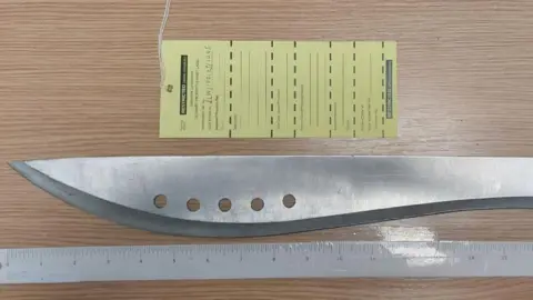 The silver of a machete on a wooden surface with a ruler next to it