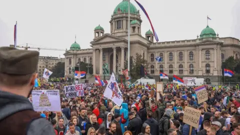 Reuters Students and anti-government demonstrators protesting in Belgrade, Serbia, on 15 March 2025. Hundreds of people holding flags and placards are gathered in front of the parliament building.