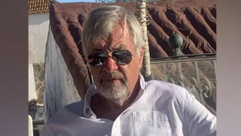A man with grey hair wearing a white shirt sits at an outdoor table with a glass of white wine. He wears sunglasses along with a bracelet on one wrist.