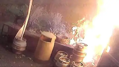 CCTV footage of the fire. There is a bin and multiple kegs stacked on top of each other. The fire is on the right side of the image and goes up to the top of the picture. 
