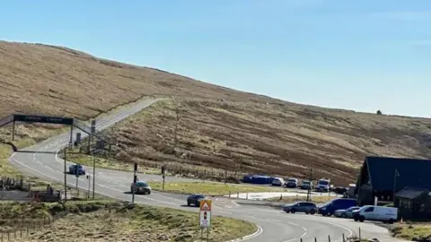 The Mountain Road stretching from the Bungalow through to Brandywell. It is on a sloping mountainside covered in heathland. There is a building at the Bungalow on the right with cars parked in front of it and there are three cars travelling on the road. There is a metal footbridge over the road on the left.