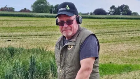 Steve Hickman standing in a green field. He is wearing a blue T-shirt with a khaki gilet over the top. He is wearing a pair of sunglasses and a baseball cap and is looking directly at the camera.