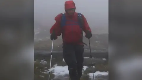 Handout Ian Evans participating in the Winter Spine Challenge in January 2023. He is wearing a red coat and walking with hiking sticks. He is along the Pennine Way and a small amount of snow can be seen on the ground.