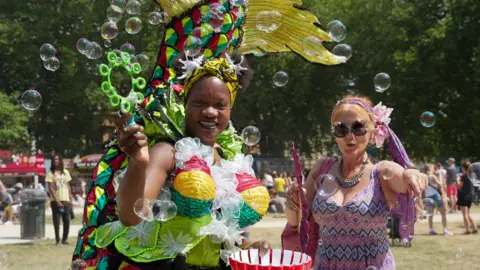 Plaster/Bristol Harbour Festival Two women in colourful costume are blowing bubbles while smiling at the camera.