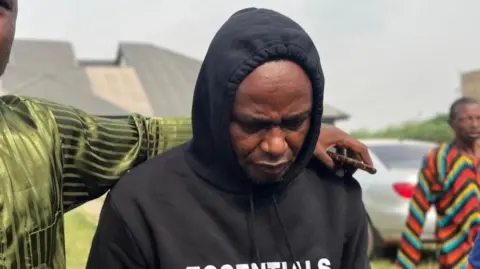 Adeniyi Kayode wears a black hoodie an looks downwards. Another man, out of shot, puts his arm round Mr Kayode