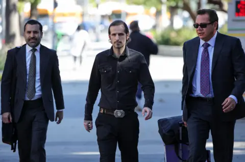 Reuters Three men outside a court wear dark clothes. Tony, in the centre, wears a dark buttoned up short, a large belt and dark trousers. He has mid length dark hair and a goatee.