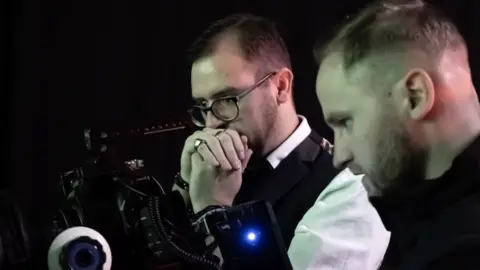 Two men in front of a black background. The man on the left has brown hair and glasses and is clasping his hands over his mouth whilst the other man is focused on a camera in front of him 