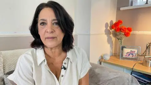 Noora sits on a beige settee in a white shirt. She has shoulder length dark hair. Ornaments and a vase of flowers can be seen behind her on a sideboard.