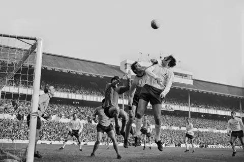 Getty Images Dozens of players gather around the goalmouth as a leaping Parkes attempt to punch a cross clear while under pressure from an opposition player. Again the stands are packed with fans in the background