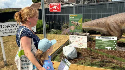 A mother and child look at a dinosaur statue which is in a metal cage. The mother is wearing a navy-blue, floral top and has a material bag over her shoulder, the girl is wearing a denim baseball cap and a light blue, floral top. The cage has several signs on it, explaining more about the dinosaur inside. 