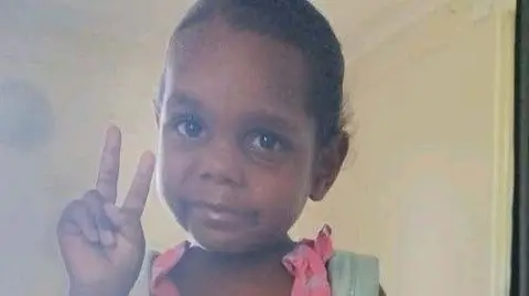 An Aboriginal girl wearing a pink dress and blue backpack poses for a picture, holding up the peace sign