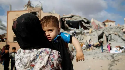 Reuters A Palestinian woman carrying a child, facing the ruins of a building in Rafah (05/05/24)