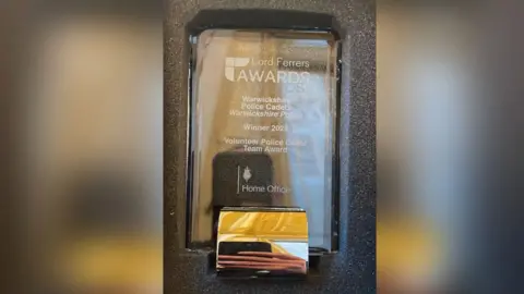 Warwickshire Police A glass trophy in a grey padded box. The inscription contains the Lord Ferrers Awards logo at the top and underneath says Warwickshire Police Cadets, Warwickshire Police, Winner 2025, Volunteer Police Cadet Team Award. The Home Office logo is inscribed below.