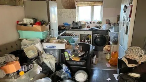 Watford Borough Council A kitchen in a flat which is littered with boxes and tupperware. Some of it is on a sofa and a chair. There is a fridge, washing machine and sink at the far end. A kitchen drawer has been pulled out. It all looks very cluttered.