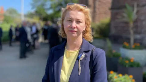 Karolina Davison, who has wavy shoulder length light coloured hair and is wearing a blue jacket over a yellow top. She is standing in the grounds of the cathedral.