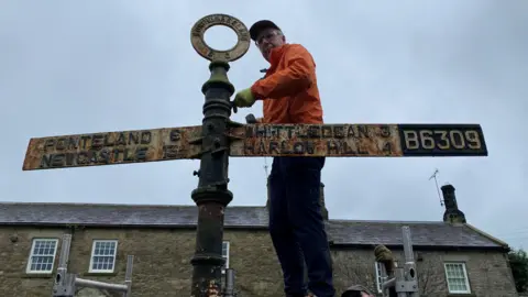 David Gosling is standing on scaffolding about 2 metres from the ground and is dismantling the same very rusty fingerpost. A row of stone terraced houses is behind him. In addition to to place names and miles, the top of the post can now be seen. It is a circle with the middle cut out - like a Polo mint - and Northumberland is written in the top half of the circle.