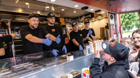 Headland Roz Eccles holds his thumbs up from inside the serving hatch of the KFC catering van as a smiling child munches through some chicken at the front of the queue.