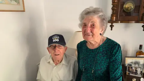 Mark Dunn Veteran Roy Booth sits in an arm chair while wearing an 100th birthday baseball cap. His wife Babs sits next to him wearing a dark green dress.