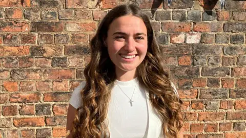 Fiona Callow/BBC A young woman dressed in white with long, curly brown hair stands in front of a brick wall. She wears a silver necklace with a cross pendant, a white t-shirt and a white waistcoat.