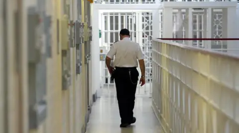 A prison officer walks down the white hall of a prison. He wears a white shirt and black trousers and has short dark hair. Prison cells are on the left side and on the right is a white metal barrier.