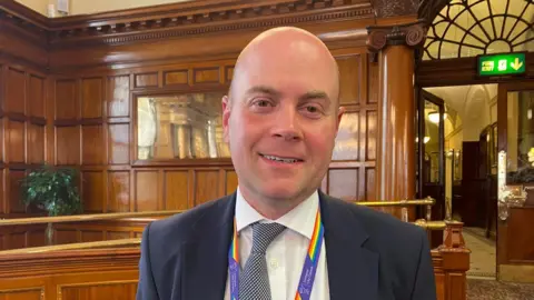 A man with a bald head smiles while he is standing inside a wood-paneled council room.
