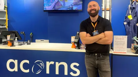 Craig Powell A man with a shaved head and a lustrous dark beard stands in front of a shop counter with a white top and a blue frontage, labelled "acorns". He is wearing a black polo shirt and black jeans and has his arms crossed in front of his body. He is smiling at the camera.