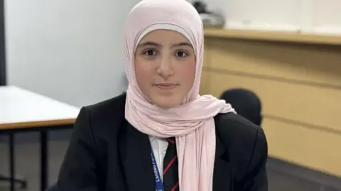 A girl wearing a pink headscarf and school blazer looks into the camera.