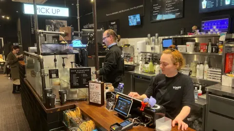 A coffee bar's tills with a server inputting an order on a screen. In the background a barista is making a coffee behind the counter in the centre of the photo. Waiting at the pick up station is a lady looking at her phone.