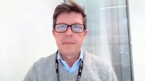 Shaun Whitmore/BBC Rory Sanderson has light brown hair and wears black rimmed glasses. He has a staff lanyard around his neck and is wearing a grey pullover and a blue shirt, which is open at the neck.