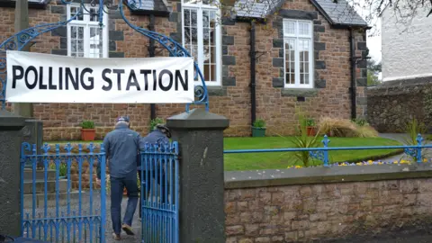 BBC Polling station sign hangs over a gateway. One of the two gates is open as two people have just finished walking through them. They are facing away from the camera - both are wearing rainproof jackets and hats.