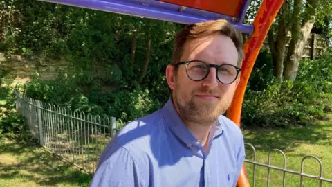A man is sitting in a park. It's a sunny day. He is wearing black glasses and a bue shirt.