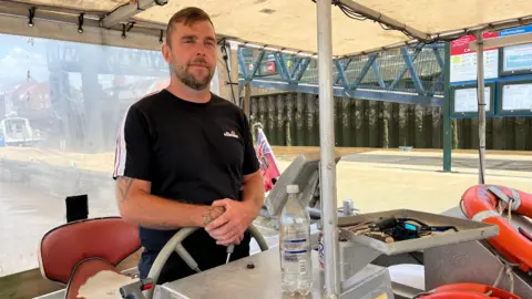 A man standing behind the steering wheel of the boat. He has short brown hair and is wearing a black T-shirt.